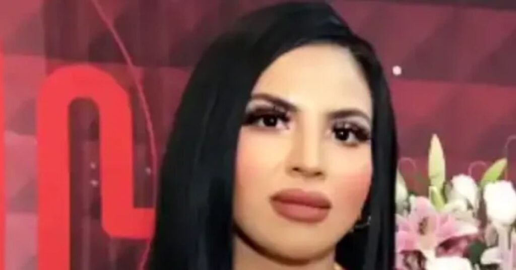 “Woman with long straight black hair and bold makeup posing in front of a red patterned background with flowers beside her.”