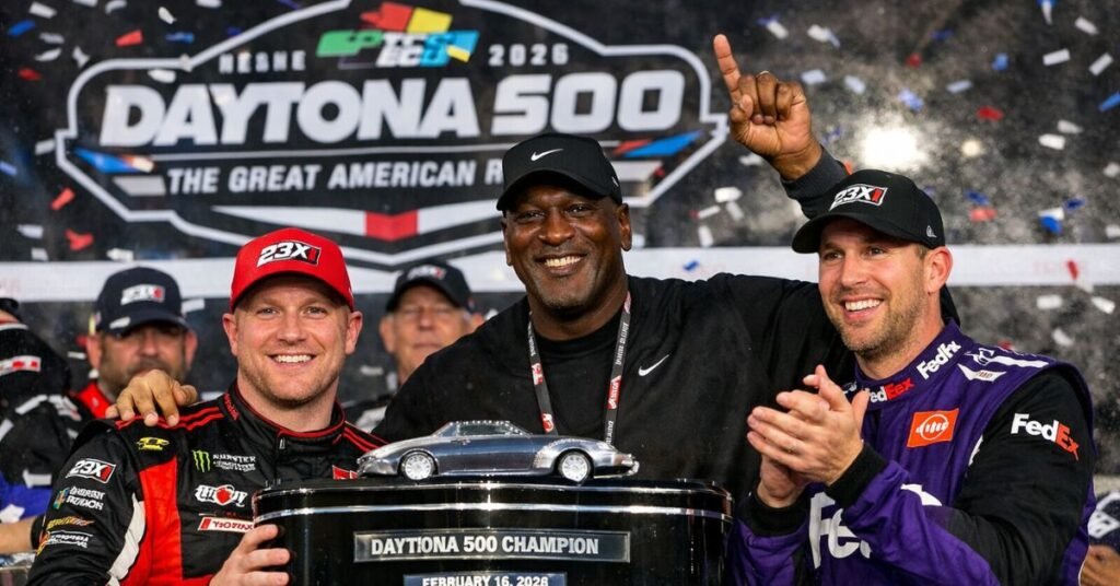 Tyler Reddick holds the Daytona 500 trophy while Michael Jordan and Denny Hamlin celebrate 23XI Racing’s victory in Victory Lane with confetti and cheering fans in the background.